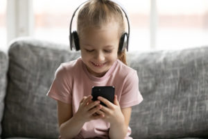 Head shot happy little kid girl wearing wireless headphones, choosing favorite music tracks in mobile application