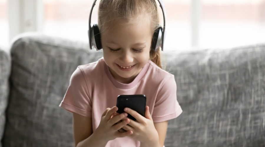 Head shot happy little kid girl wearing wireless headphones, choosing favorite music tracks in mobile application